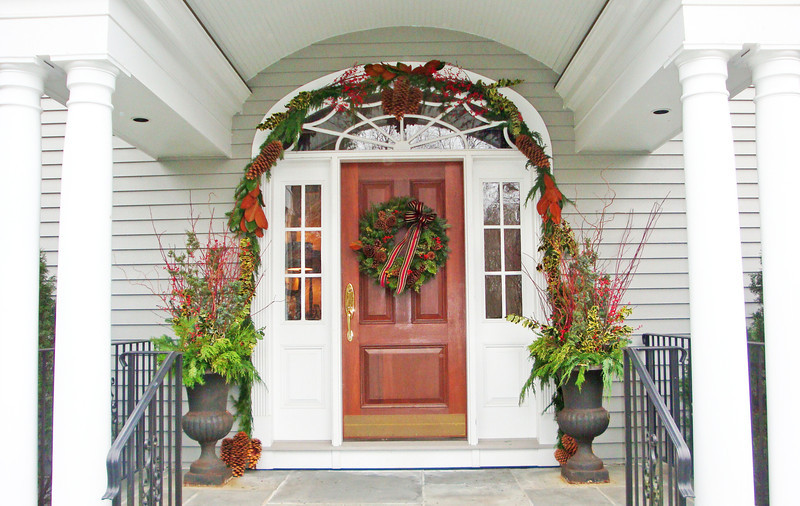 wreath with a bow on exterior door, framed by garland wrapped around door frame with Christmas arrangements in planters